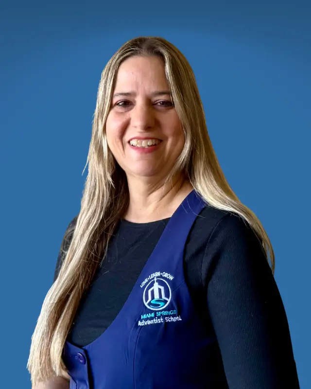 Smiling woman with long blond hair wearing a navy blue school apron with a logo, standing against a blue background.