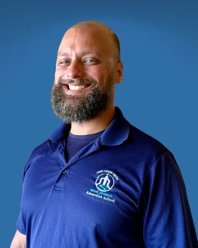 Smiling man with a beard wearing a navy polo shirt with a school logo, standing against a blue background.