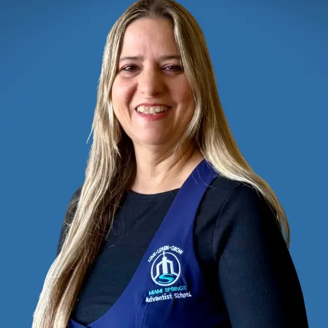 Smiling woman with long blond hair wearing a navy blue school apron with a logo, standing against a blue background.